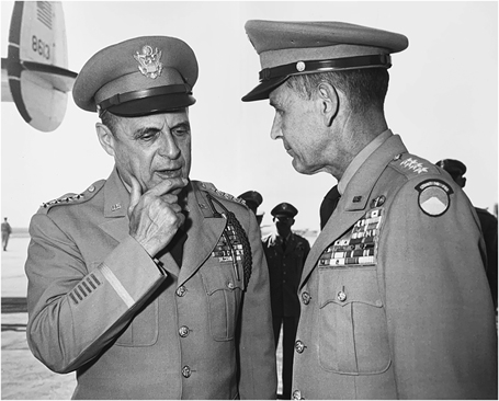 Figure 4. Two generals stand together in discussion on an airfield. The general at left has his hand on his chin. Additional military personnel can be seen in the background, along with the tail of an aircraft.