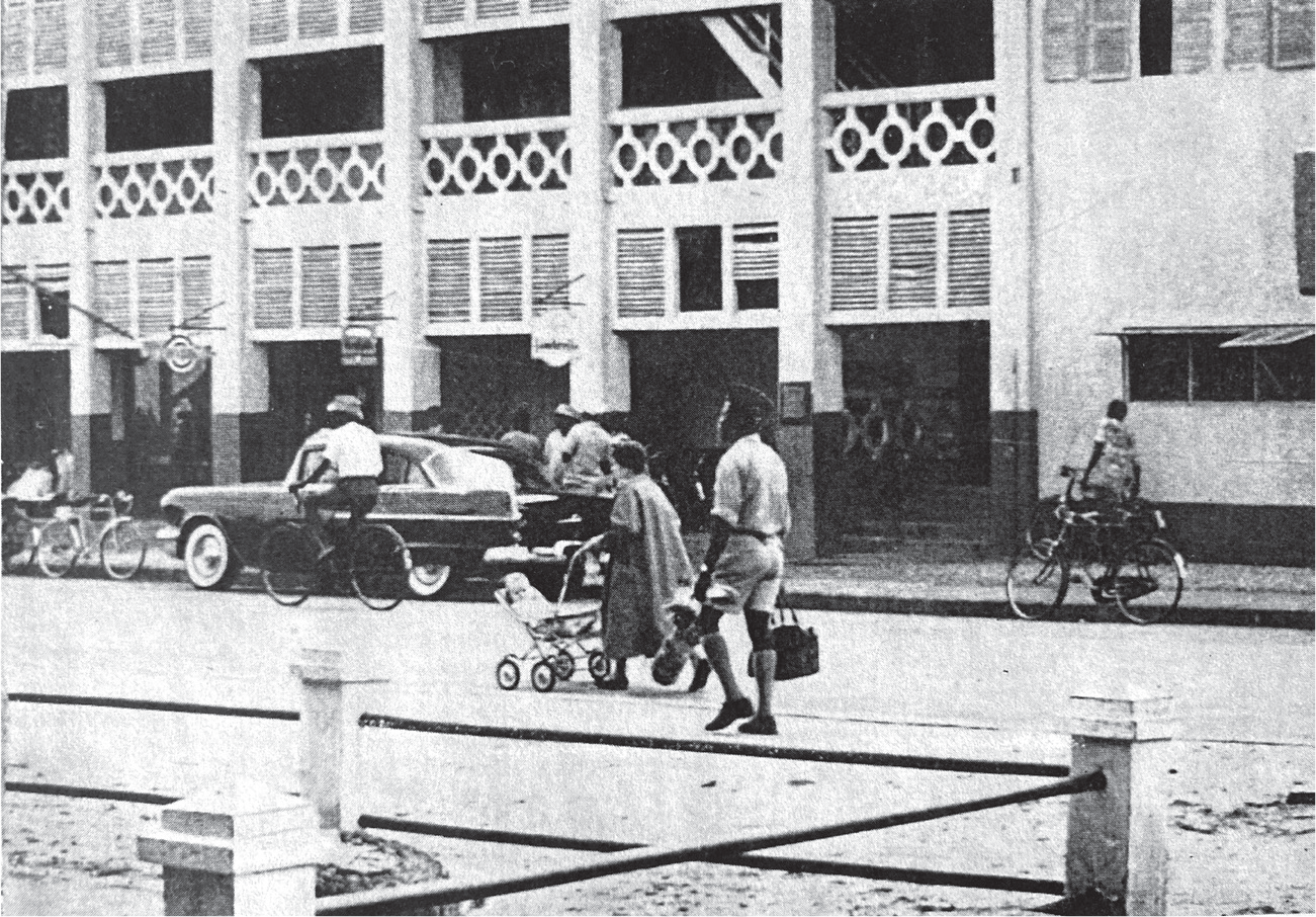 A white woman pushes her baby carriage while her African servant carries her bags several feet behind in Cotonou, Dahomey.