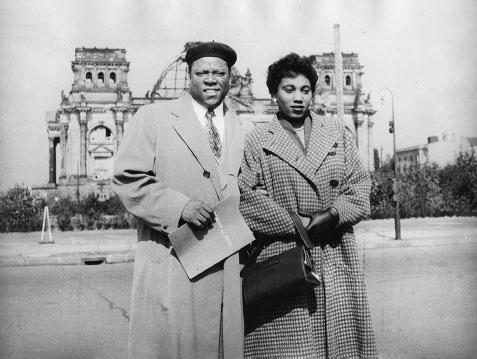 A Black man and woman stand outside wearing warm-weather coats. Behind them is the bombed-out German Reichstag building.