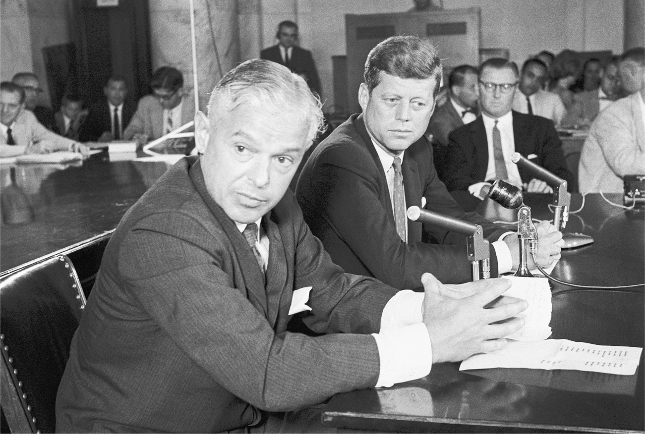 Two well-dressed men sitting in front of microphones. Nitze is in the foreground and is speaking to an unseen person out of the frame to the camera’s left. Kennedy is sitting to Nitze’s left, looking contemplative.