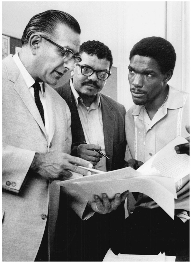 A white man gestures at papers in his hand to two Black men. One of them is looking where he is pointing while the other is looking at him intently.