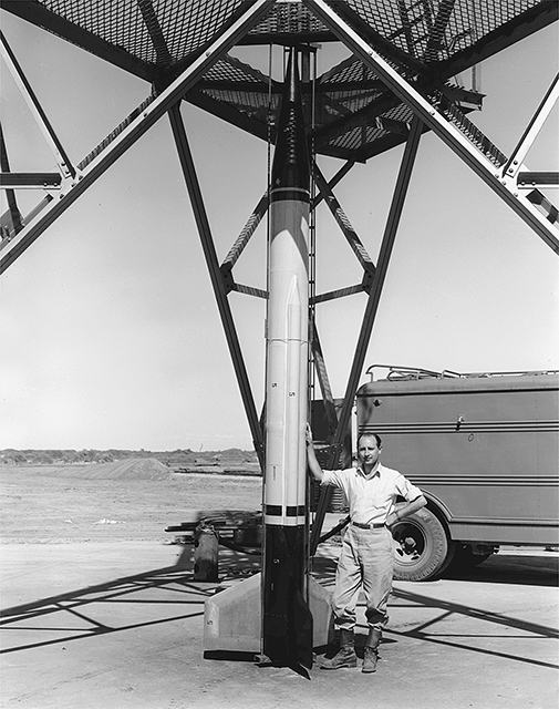 A man wearing a shirt with the sleeves rolled up and tall boots poses stands under the large metal framework of a rocket.