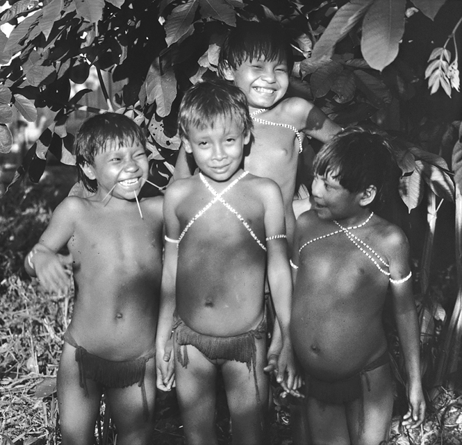 Four Yanomami children smile and laugh. Three have white necklaces looped around their necks and onto their upper chests and around their upper arms. One child has three sticks protruding from their lower jaw.