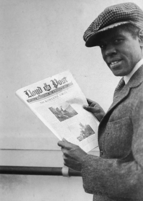 A Black man wearing a fashionable cap and a wool suit smiling at the camera holds up a copy of a newspaper with the headline, “THE ROMANTIC RHINE.”