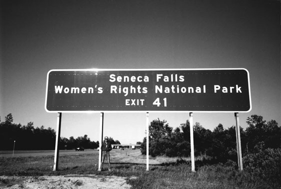 Figure 25 An unusually large interstate highway sign, requiring five support posts because of its length, reads “Women’s Rights National Park,” all on one line. The sign dwarfs a man standing below it.