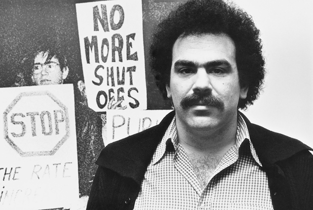 Figure 51. A man stands in front of a large photo of people with signs that say, “No More Shut Offs” and “Stop the Rate Increase.” He has bushy hair and a dark mustache. He stares unsmiling at the camera.