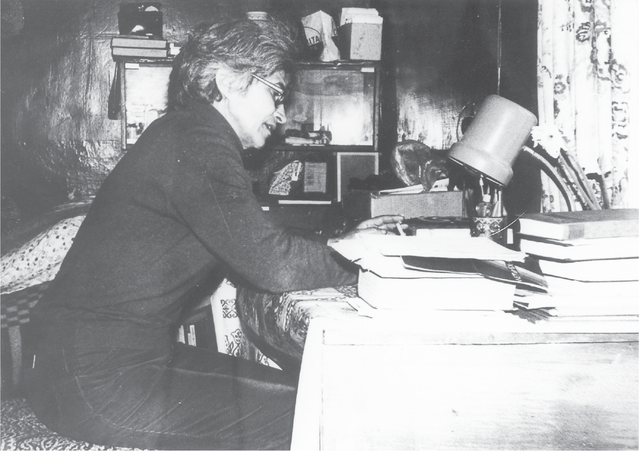 In profile, a thin, late middle-aged woman in glasses sits at a desk crowded with books and papers.