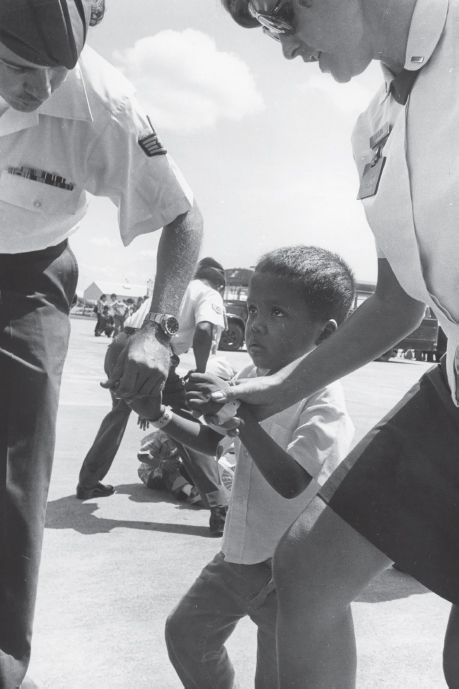 Two US Air Force service members, one male and one female, assist a South Vietnamese orphan from a plane at Clark Air Force Base in the Philippines. The young boy appears to be of mixed race. Another service member helping a child off the ground is seen in the background.