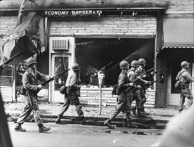 Figure 11. A platoon of six soldiers walks down a devasted street, bayonets fixed on rifles that they hold at the ready as they pass the Economy Barber Shop, its glass window shattered, the street littered with debris.