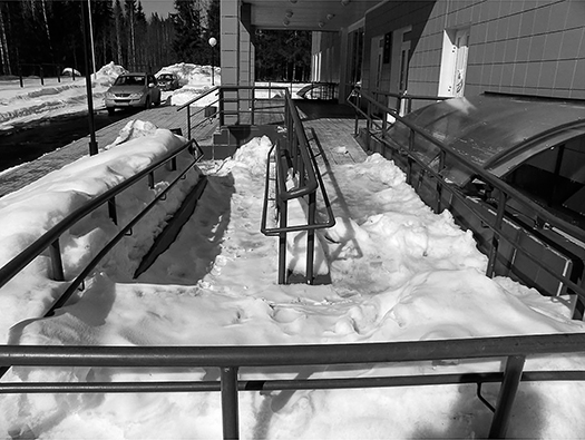 Snow covers an accessibility ramp with handrails. The steps to the front entrance of the building have been shoveled, but the ramp has not. Footprints show that some visitors have used the ramp anyway, but the deep-drifted snow along parts of the ramp would make it difficult for a wheelchair user or someone pushing a stroller to navigate.