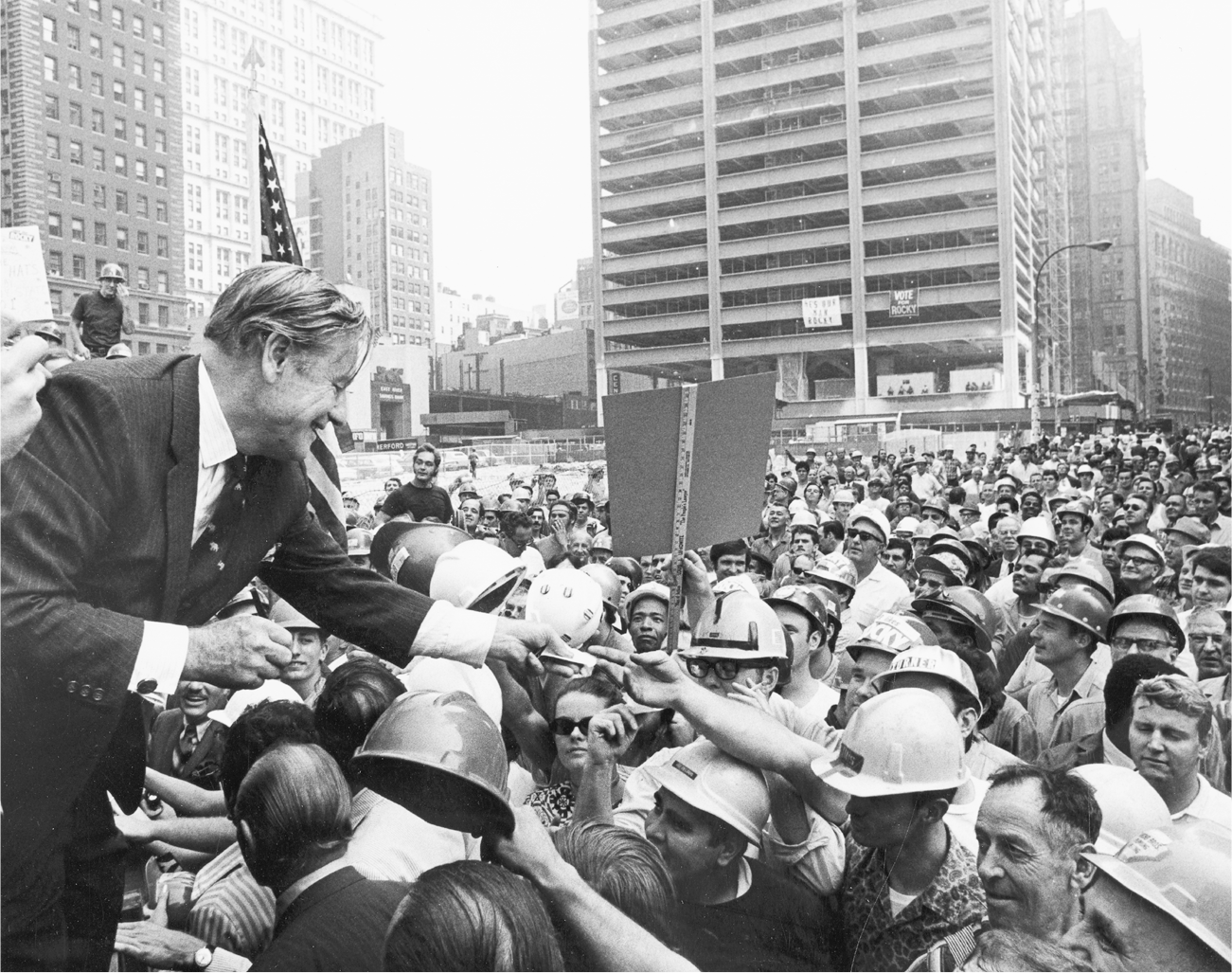 Rockefeller signs autographs from a raised platform at a construction site. The autograph seekers are largely male workers wearing hard hats. Some have pro-Rockefeller stickers affixed to their hats.
