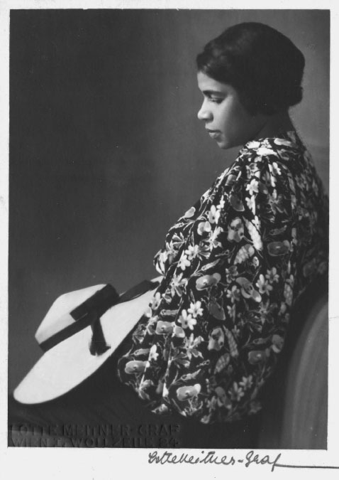 A young Black woman wearing a floral print shirt and black slacks stares down at the fashionable straw hat with a black ribbon wrapped around it in her lap.