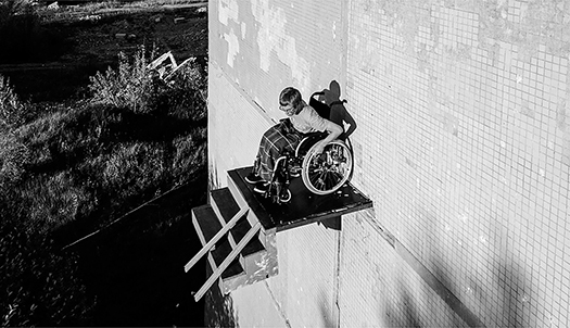 Another angle of the ramp high on the side of a building, taken from slightly above. The man sits with his hands on the wheel guides, leaning forward and looking over the edge of the platform, as if wondering where to go, or contemplating the lack of survivable options the ramp offers.