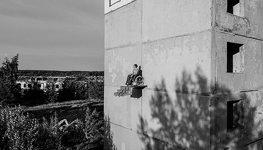 A wide view of a man in a manual wheelchair wearing pajama pants and a t-shirt who sits on a platform suspended three stories above the ground overlooking treetops, a field, and nearby buildings. He looks pensively into the distance and sits with his hands in his lap.