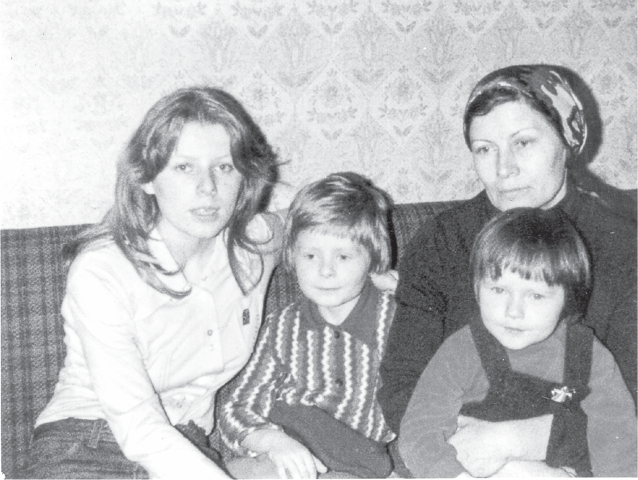 A mother, looking preoccupied and sad, with three children on a plaid sofa. The daughter on the left, looking serious, has her arm around the back of a young boy as the mother holds a little girl.