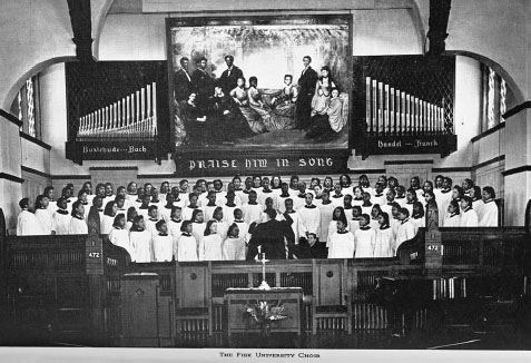 A choir of approximately fifty young men and women dressed in white robes stands inside the front of a church hall, looking towards the conductor, a tall man in a black suit standing before them.
