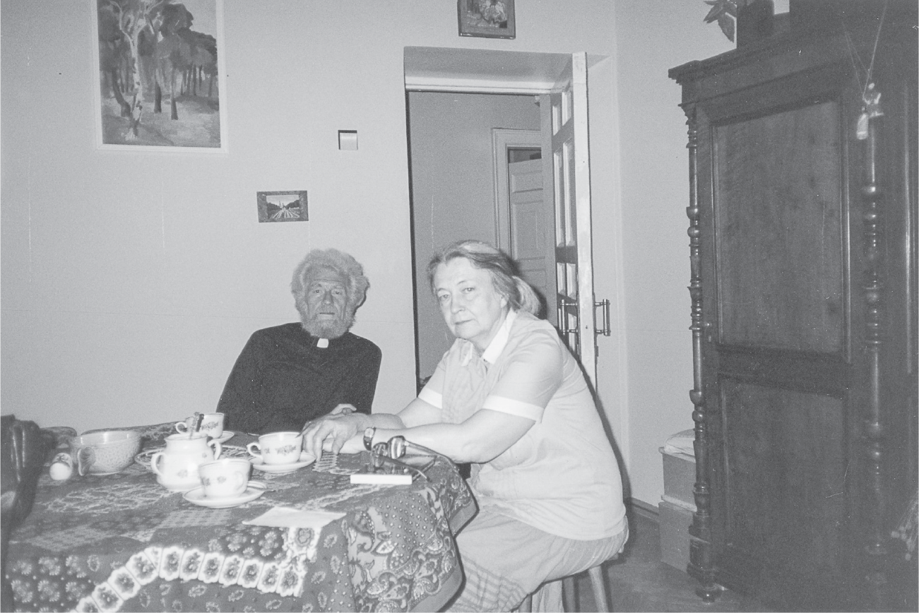 A man in a white priest’s collar with thick white hair and a full beard and a gray-haired woman look serious. They sit at a circular table with a vibrant patterned tablecloth and teacups.
