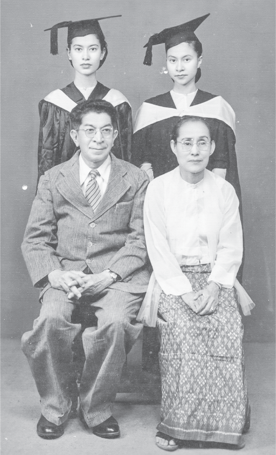 Auntie Rosie and her sister Jenny, looking serious in graduation caps and gowns, pose for a studio photo with their mother and father, seated in front of their daughters and dressed, respectively, in a sheer muslin blouse, shawl, and silk longyi, and a suit and tie.