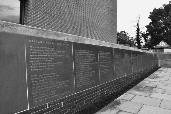 Figure 21 An even row of a dozen or more dark engraved stone tablets resembling printed pages of a book extend across a plaza with stone paving at their base and a brick wall rising behind.