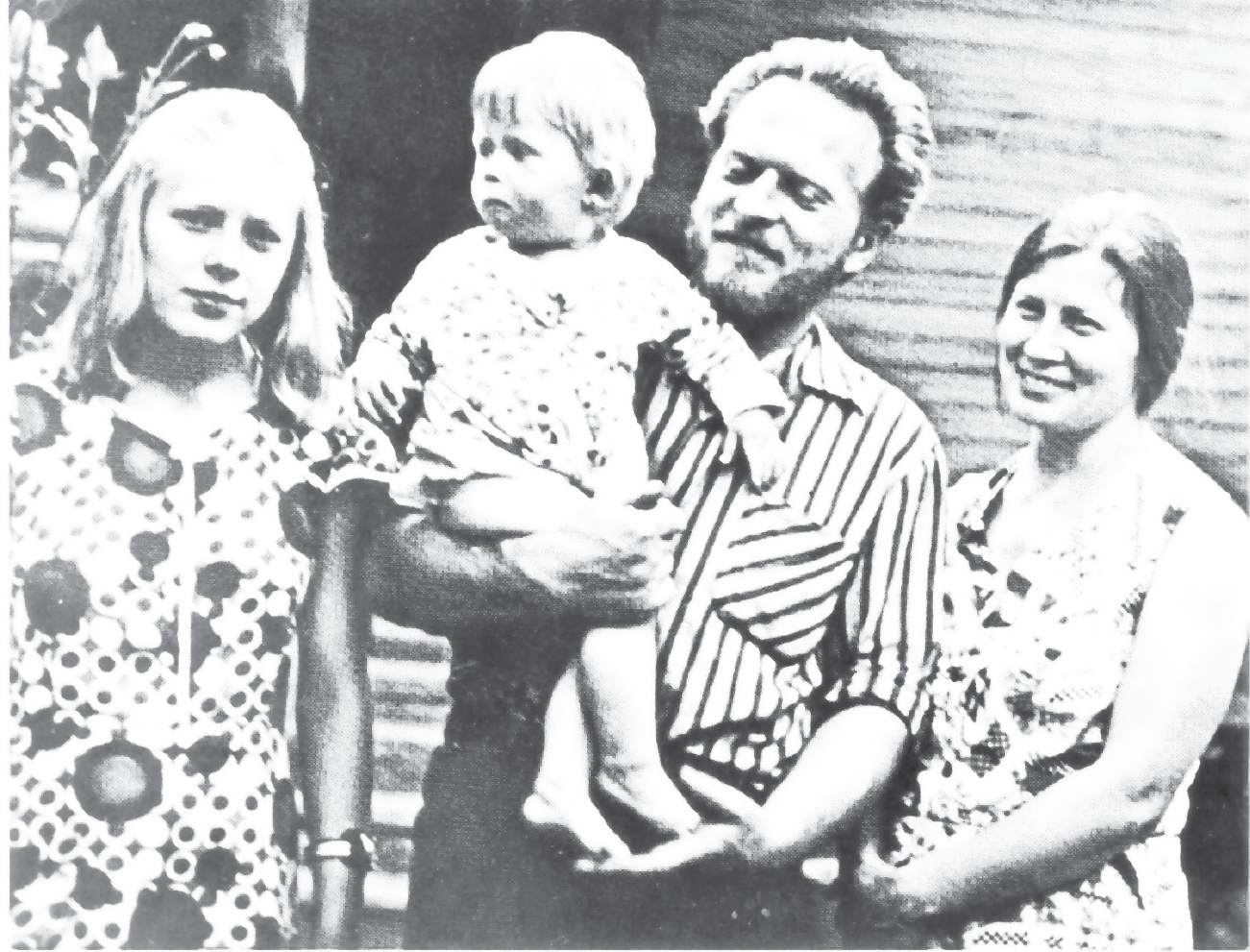 A young family of four. The light-haired adolescent daughter on the left looks thoughtful. The father and mother smile happily as the father (center) holds a light-haired toddler who looks serious, his head turned in the direction of his sister toward something outside the picture.