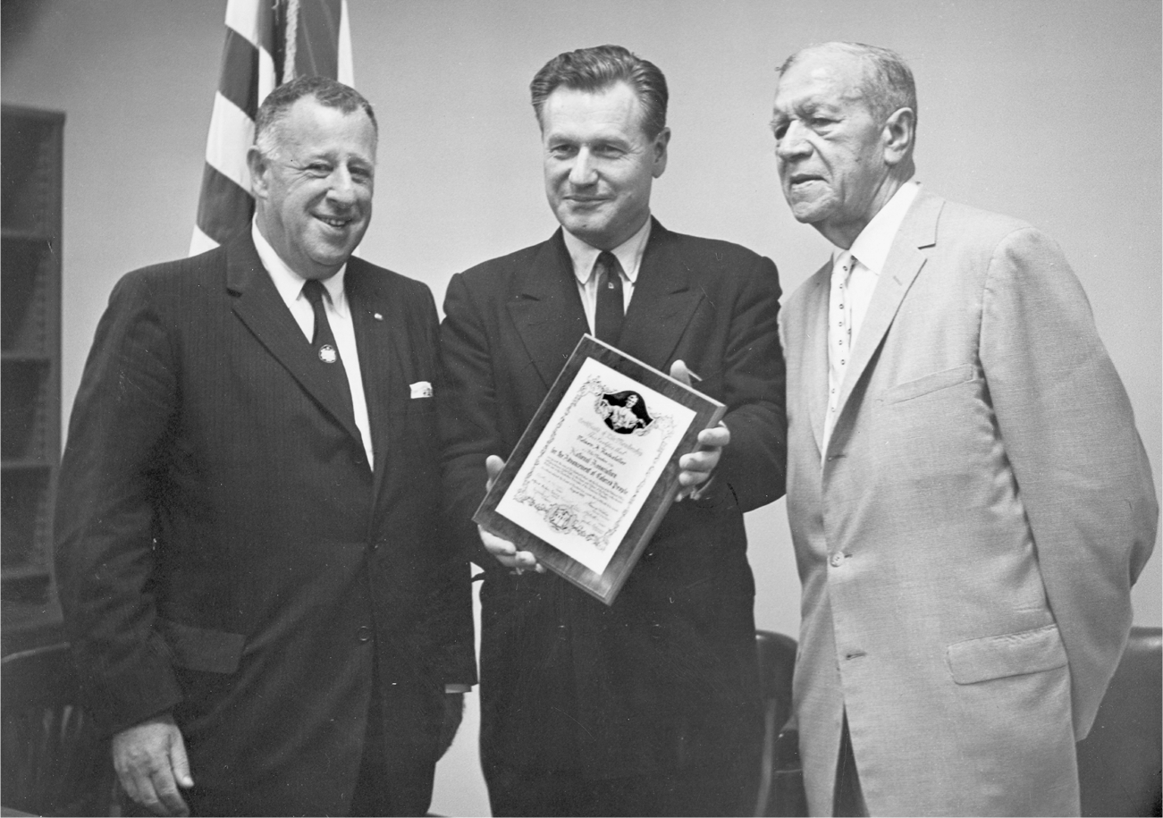 Rockefeller poses for a photograph in between two other men. With a proud look on his face, he holds a plaque that he displays to the camera indicating that he is now a life member of the National Association for the Advancement of Colored People.
