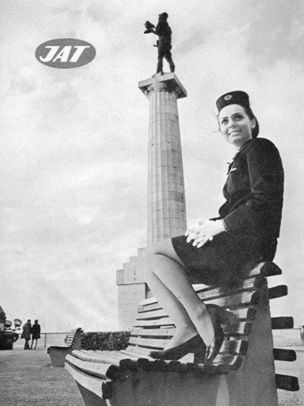 Figure 8.5. A stewardess in a cap, blazer, skirt, and pumps sits on top of a bench, her feet resting on the bench’s seat. She smiles and looks above the camera. A tall monument is behind her, and the “JAT” logo appears in the top left of the image.