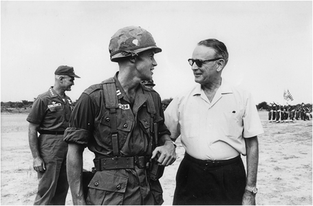 Figure 9. Two men, one in military gear and the other in civilian clothes, have a conversation outside, as troops on parade can be seen in the distance. A smiling general in fatigues is visible behind them.