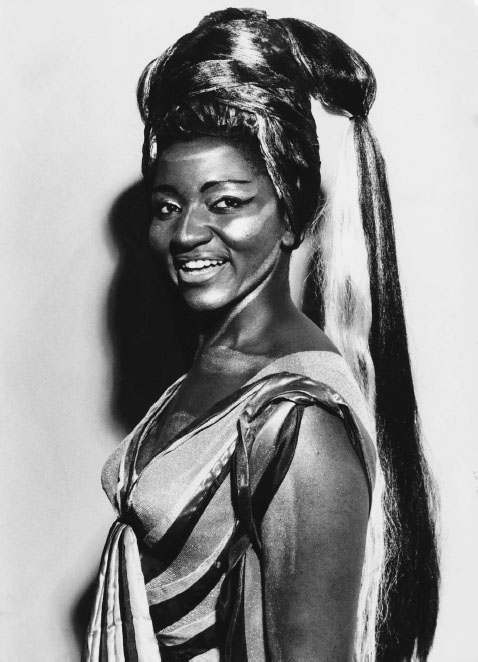 A Black woman wearing a long wig and gold makeup on her face smiles for a studio portrait photo.
