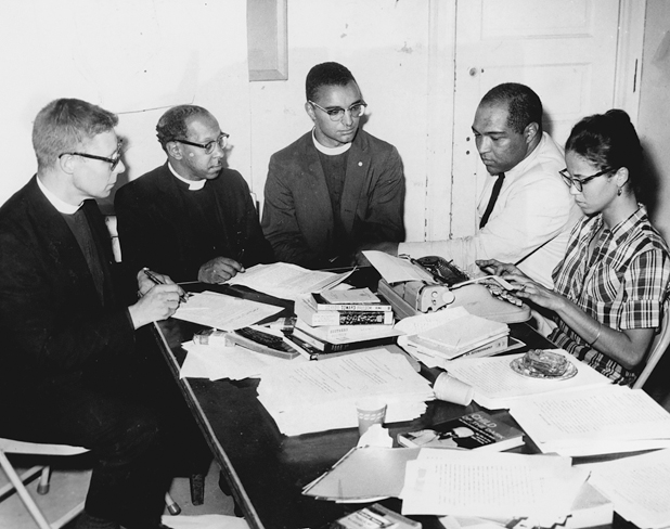 Figure 2. A group of five people are seated around a table filled with papers and books. There are three clergymen, wearing their religious collars, one young woman typing and one man in shirt sleeves. All are looking intent and focused.