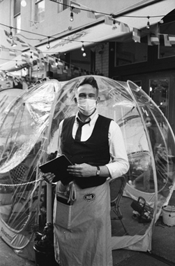 Figure 17. A masked waiter stands outside a restaurant’s clear plastic dining enclosure on the Upper West Side of Manhattan.
