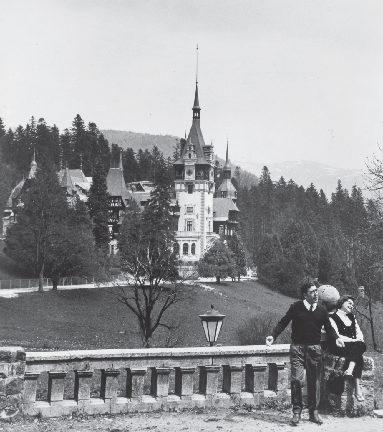 A man and woman lean against against a stone bridge at the bottom right of the picture. There is a castle in the distance surrounded by large trees. Mountains appear behind the castle.