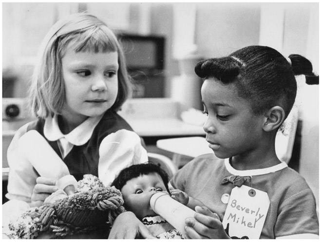 A young white girl gazes at a young Black girl as both pretend to feed bottles to the baby dolls they are holding. The Black girl has a name tag that reads “Beverly Mikel.”