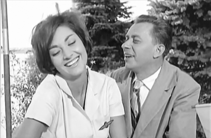 Figure 8.2. In a frame from the film Love and Fashion, a man in a light-colored suit and a patterned tie gazes at a stewardess in a white blouse while both laugh. Evergreen trees are visible behind the pair.