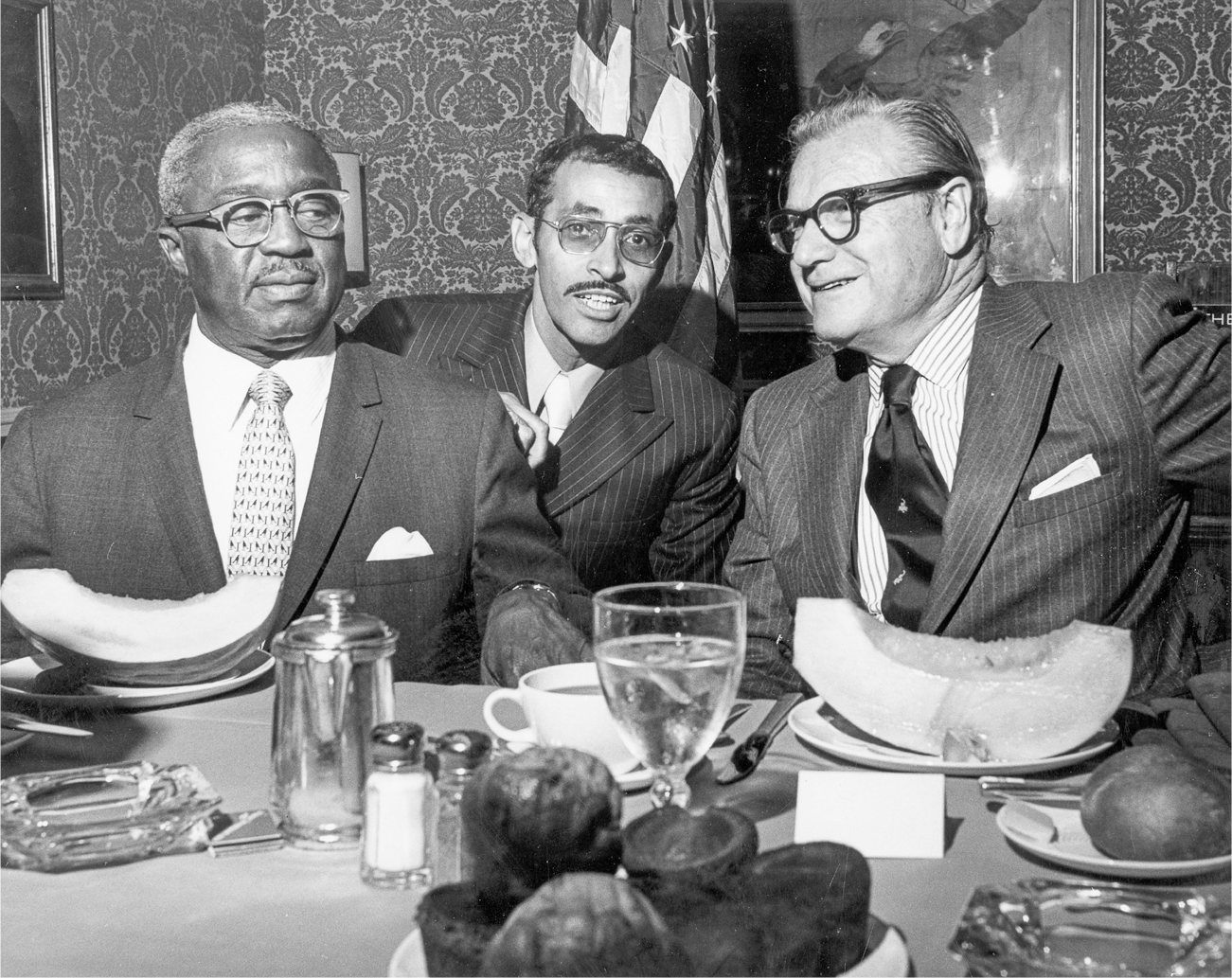 Rockefeller sits at a table set for dinner alongside two men. They all wear suits with an American flag hanging from a pole behind them.