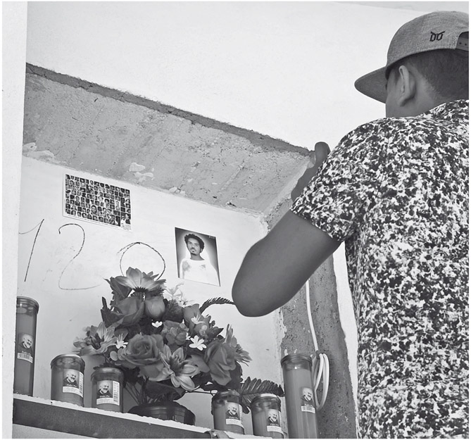 Figure 17. A young man wearing a baseball cap takes a picture with a mobile phone of a grave placed in a multistory construction. The number 120 is written on the concrete surface and next to the number is a photograph of a young man and a postcard depicting a grid of faces. There are flowers and candles placed on the sill.