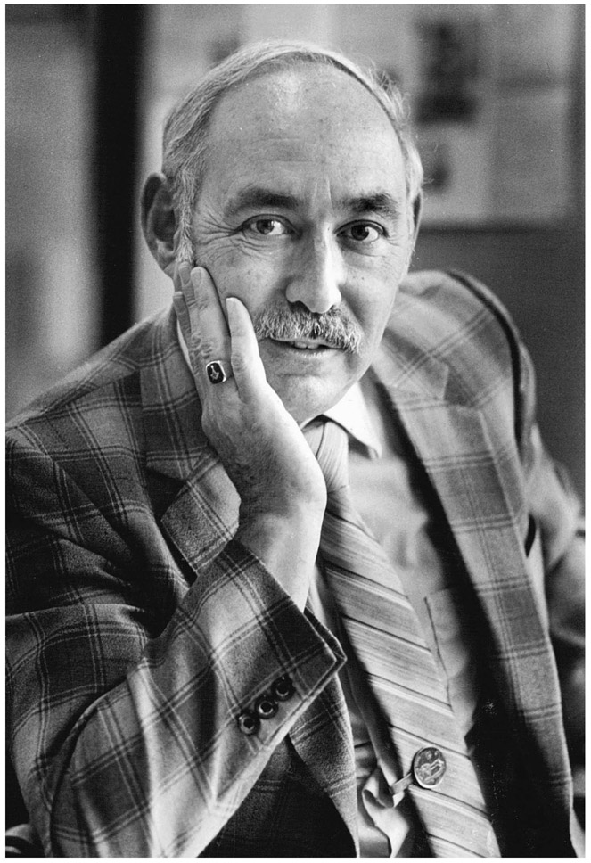  A man in a checkered sports coat and striped tie poses while seated with his chin in his right hand.