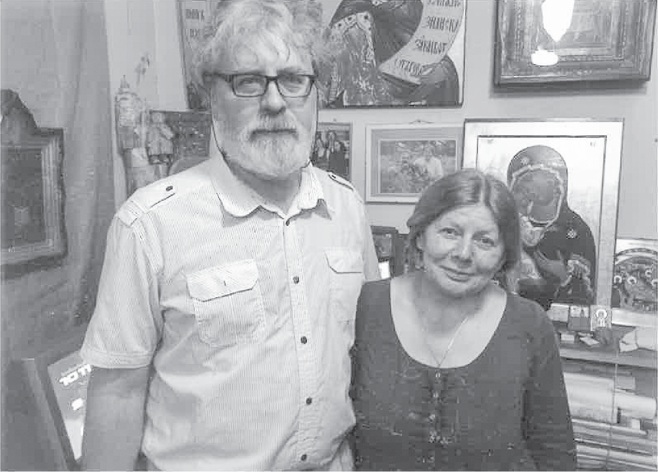 A middle-aged couple stands in front of a wall of icons. The man wears glasses and has a white beard and white hair. The woman, smiling slightly, comes up to his shoulder and has pulled-back middle-parted brown hair.