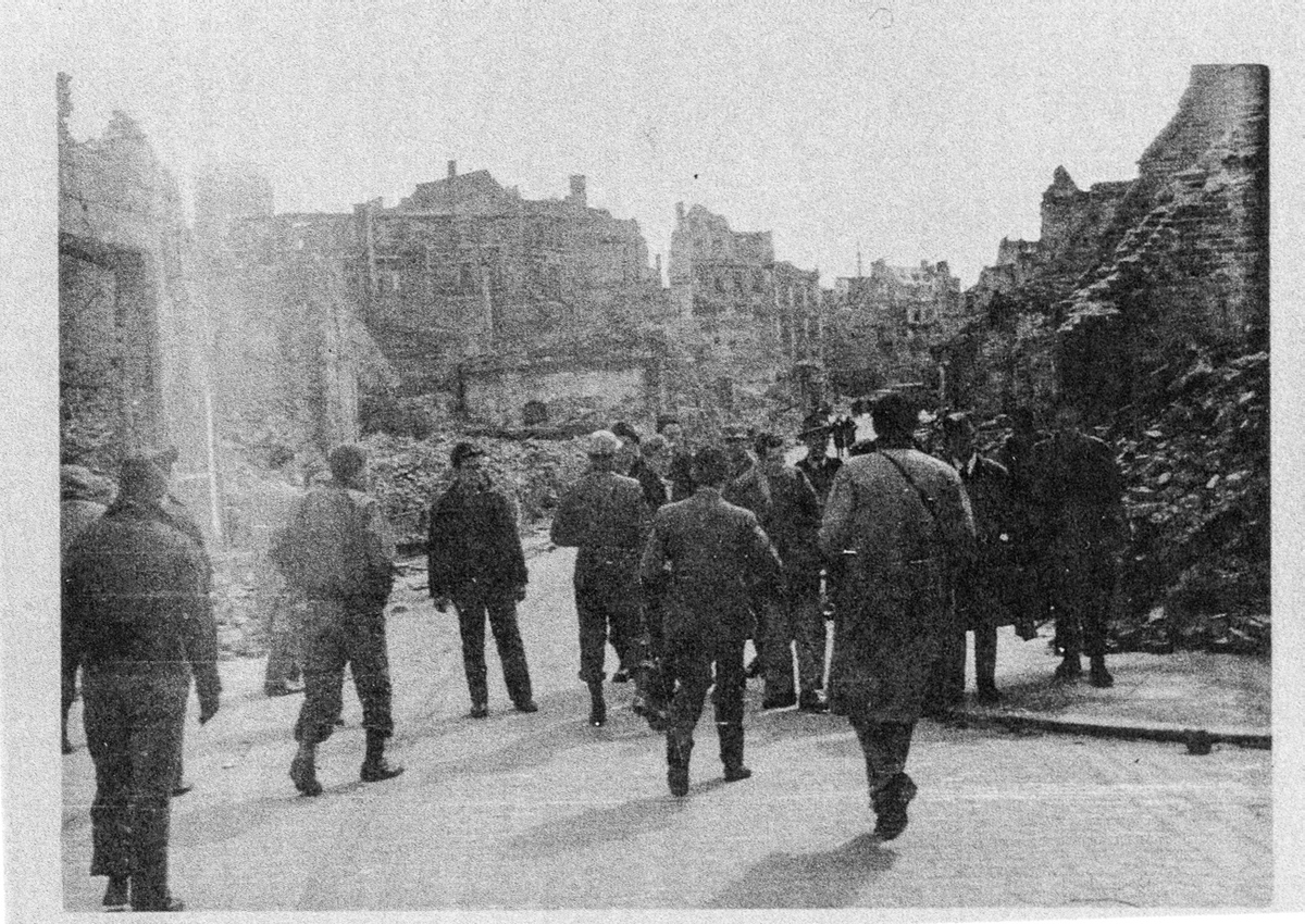 Figure 6. A group of men walk together; those in the forefront walk away from the camera and toward a group facing the camera. In the background is the ruins of a city.