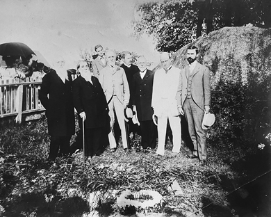 Seven men of prominence, mostly elderly, all nicely dressed and a few in light summer suits, stand by a grave. The grave itself is mounded high with wreaths.