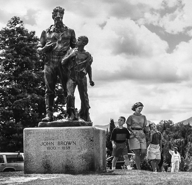 A woman and two children walk past the bronze statue of John Brown and a Black boy, anchoring a grassy traffic circle at the John Brown Farm in North Elba.