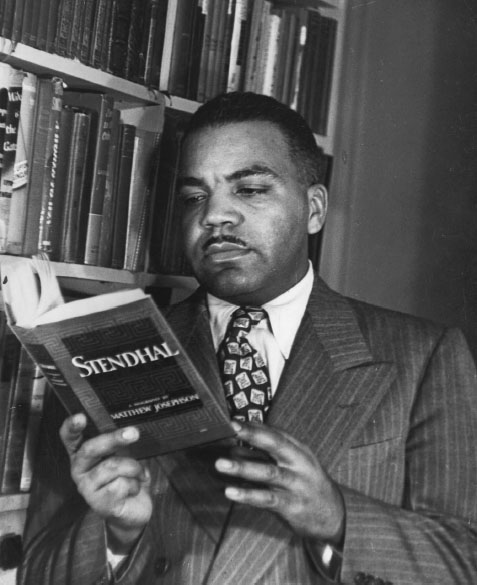 A Black man standing next to a bookcase and wearing a suit stares down in concentration at the book in his hands.