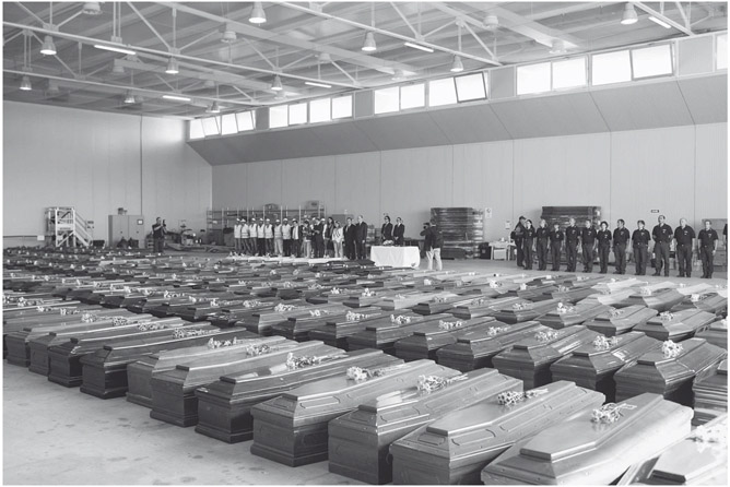 Figure 3. The floor of a large hall is covered by coffins with flowers on top of each one. Two groups of ten to fifteen people stand in rows in the back of the hall facing the coffins.