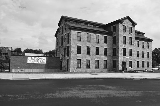 Figure 24 A four-story mill building of massive cut-stone blocks and well-maintained appearance adjoins a brick wall with a banner reading “National Women’s Hall of Fame.”