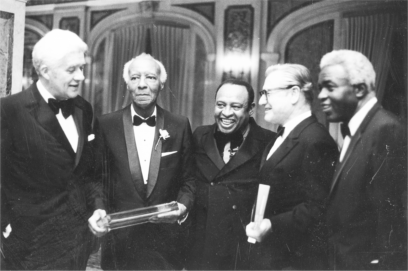 A group of five men, including Rockefeller, stand in tuxedos as they appear to enjoy each other’s company. Two of them hold glass mementos.