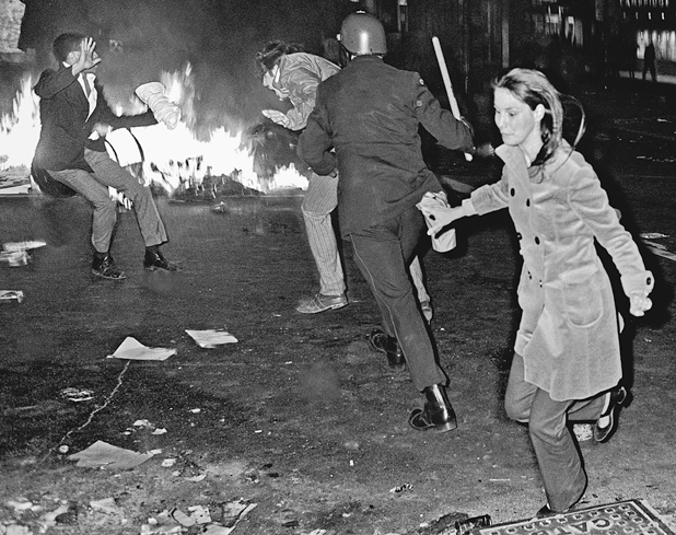 Figure 39. A policeman charges two protestors his baton ready to hit them while a young woman runs in another direction. In the background fires are burning in the street.