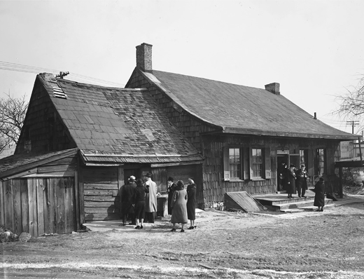 Figure 3.4. A photo of a modest one-story wood-frame house with an extension and shed includes ten white men and women who appear to be inspecting the building. The roof of the extension needs repair, and the space in front of the house is not landscaped.