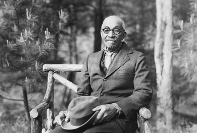 Lyman Epps Jr, the last North Elban with a connection to the Black pioneers, poses in a wide rustic chair, his good felt hat in his lap. Pine trees rise behind him, and the strong light of the sun shows the whiskers of his mustache and the faint hint of a beard.