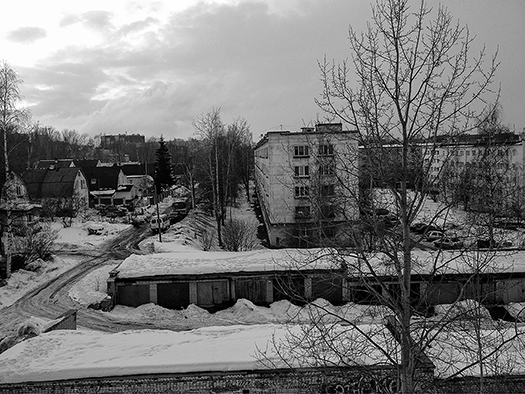 A block of five-story apartment buildings wraps around a snowy yard with parked cars. A row of garages with doors is painted in various colors. Several stand-alone houses are depicted. Small birch and evergreen trees pepper the landscape. The sky is low and gray, with sunlight coming in from the side.