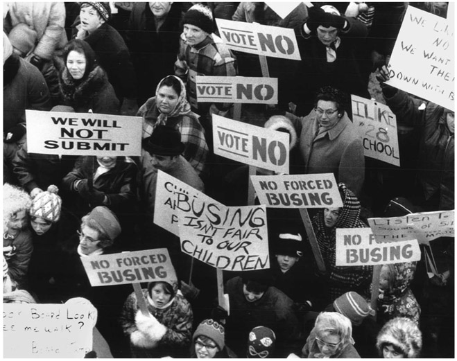 A portion of a crowd of white people seen from above, dressed in winter clothing and holding signs, including: “WE WILL NOT SUBMIT; NO FORCED BUSING; BUSING ISN’T FAIR TO OUR CHILDREN; VOTE NO.”