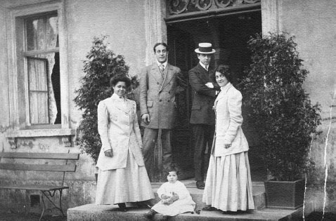 An African American woman stands outside on the steps of an apartment building with two adult white men in suits and a white woman to her right. On the step next to her feet is a small child dressed in white.